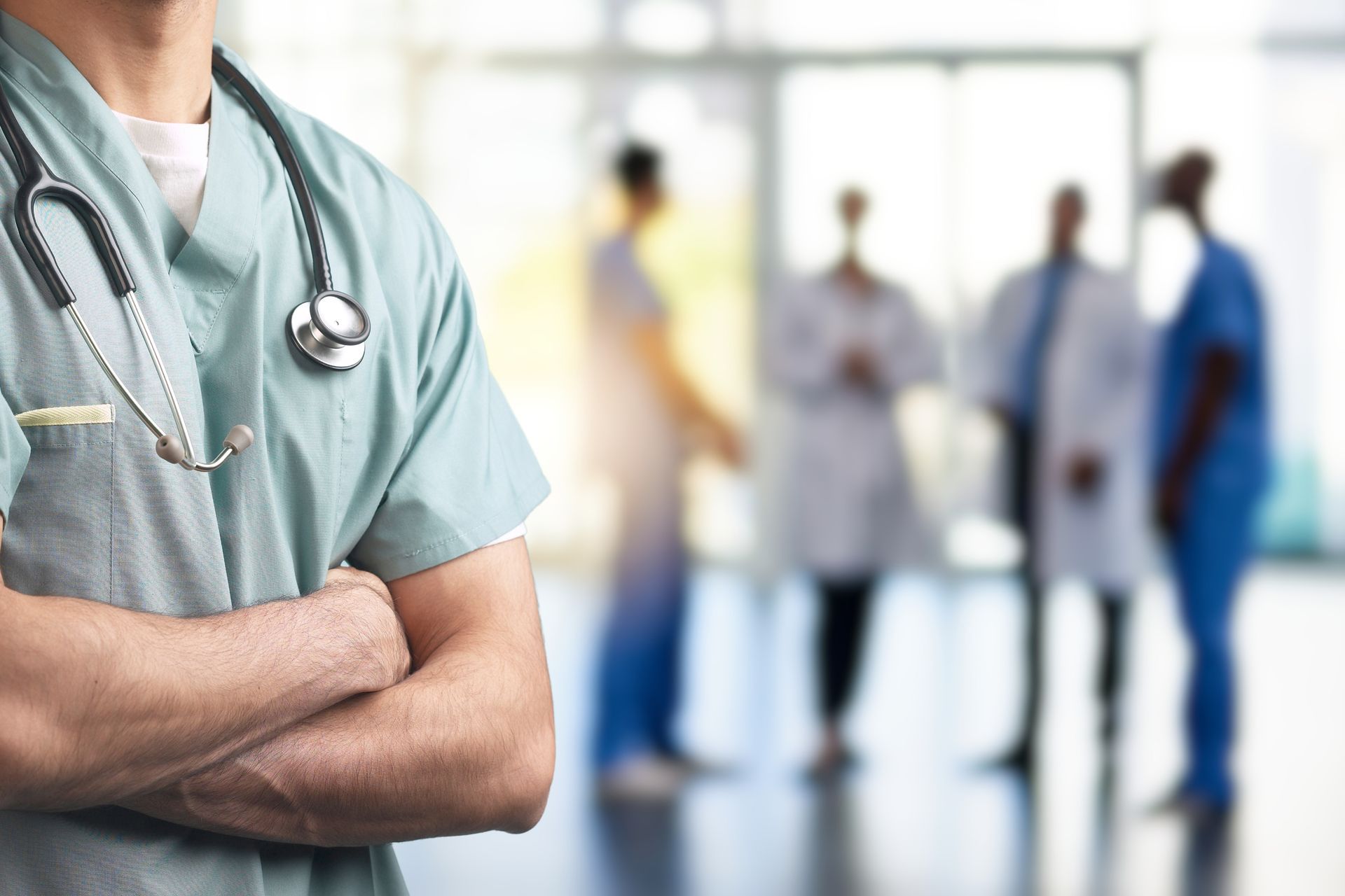 A doctor with a stethoscope around his neck is standing in a hospital with his arms crossed.