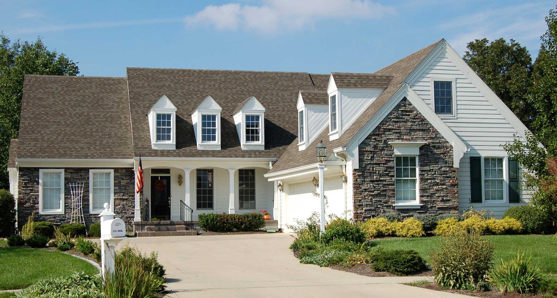 Suburban house with stone facade, white siding, front porch, driveway, and manicured garden under blue sky