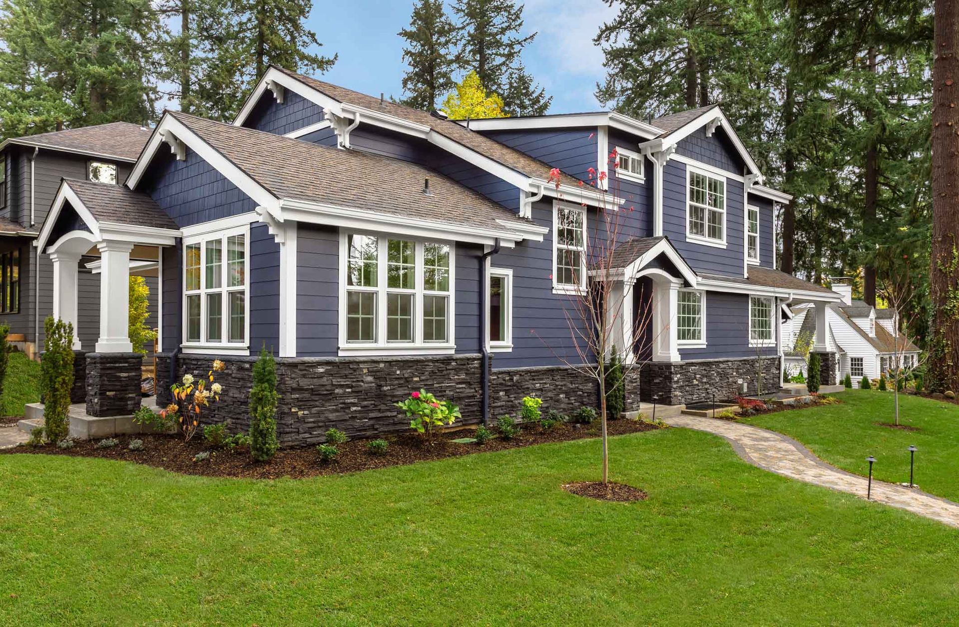 Gray two-story house with stone accents and a manicured green lawn amid tall trees