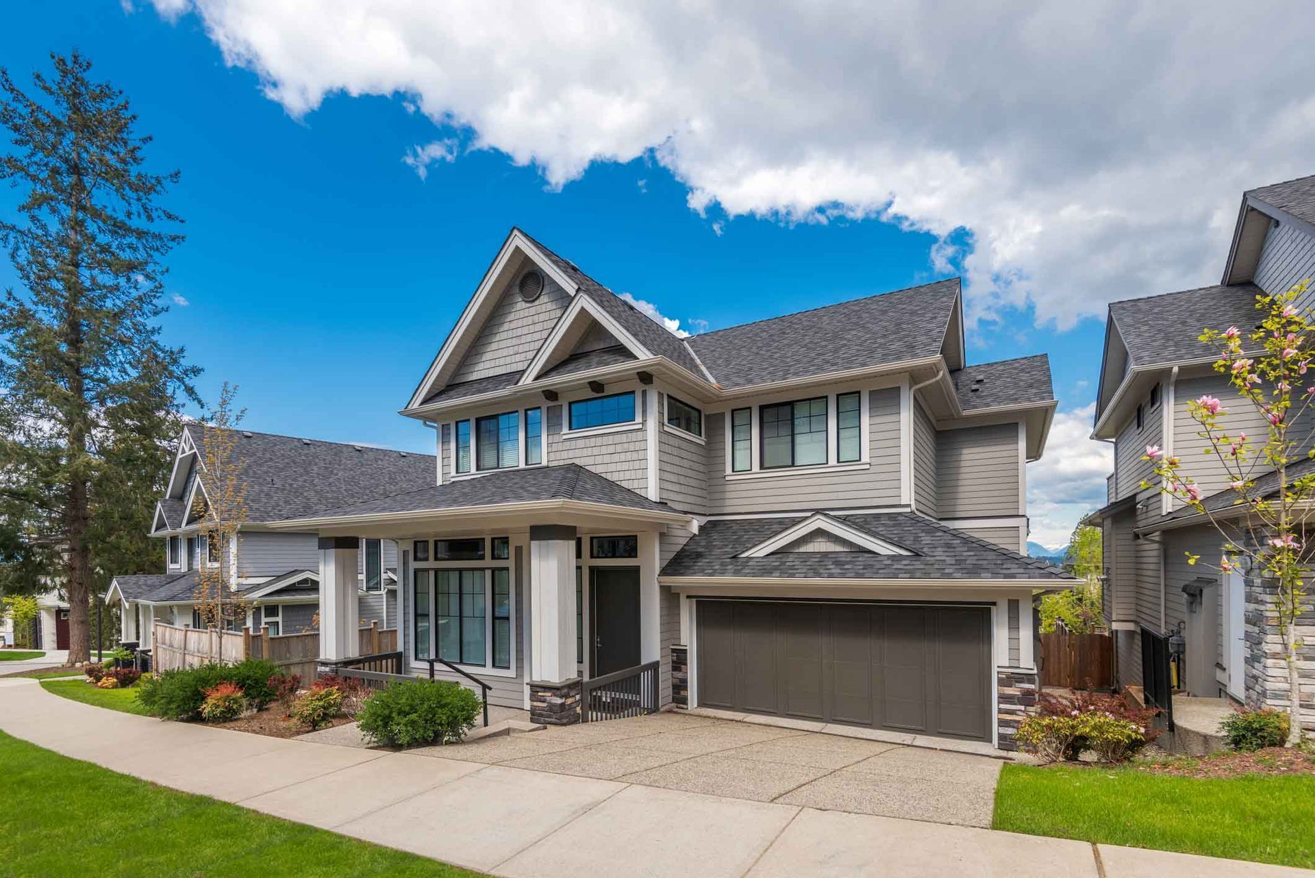 Two-story gray suburban house with a front porch, garage, and landscaped lawn under a blue sky