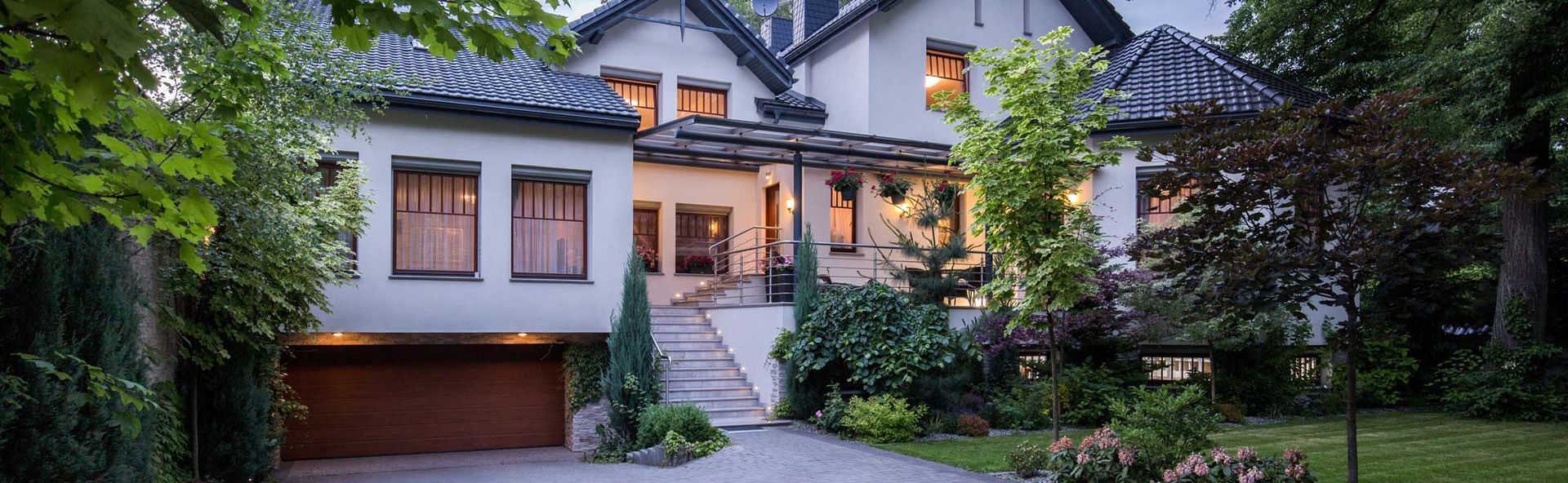 Modern two-story house with a lit entrance, garage, and lush green landscaping at dusk