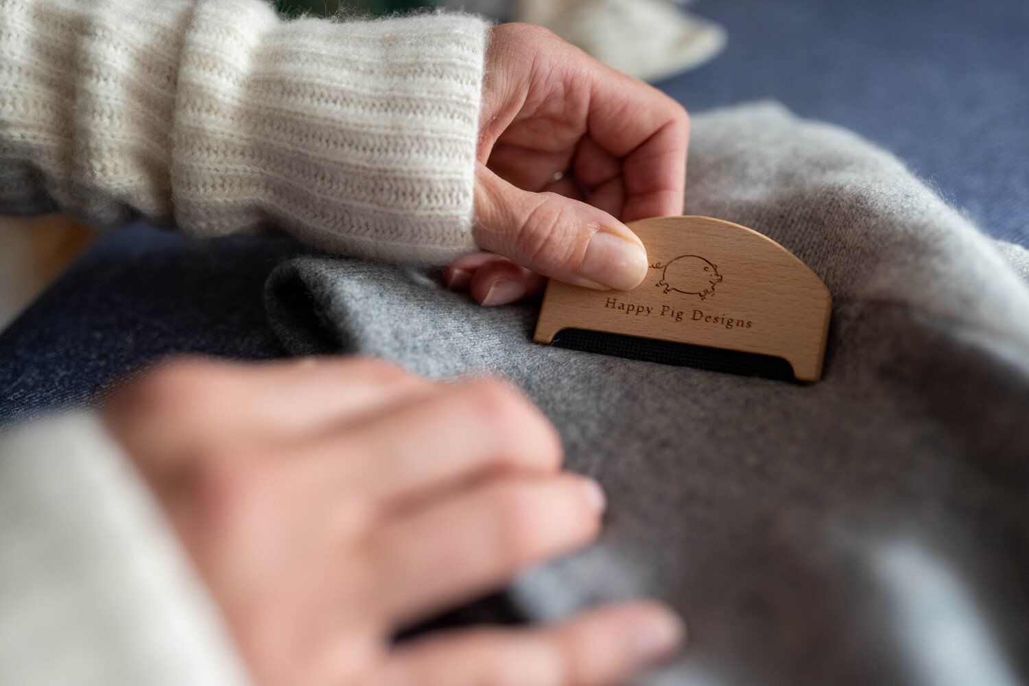 a hand holding a wooden cashmere depilling comb against a jumper. the comb has a logo with a pig on it
