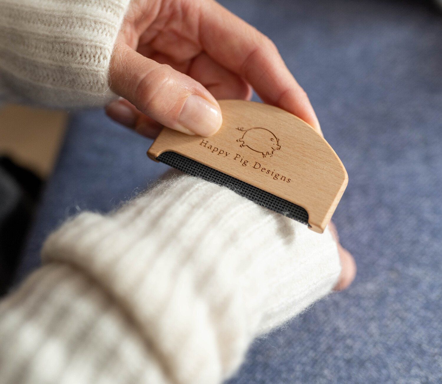 A photo of a hand holding a cashmere depilling comb with the logo Happy Pig Designs on it against a cashmere sleeve