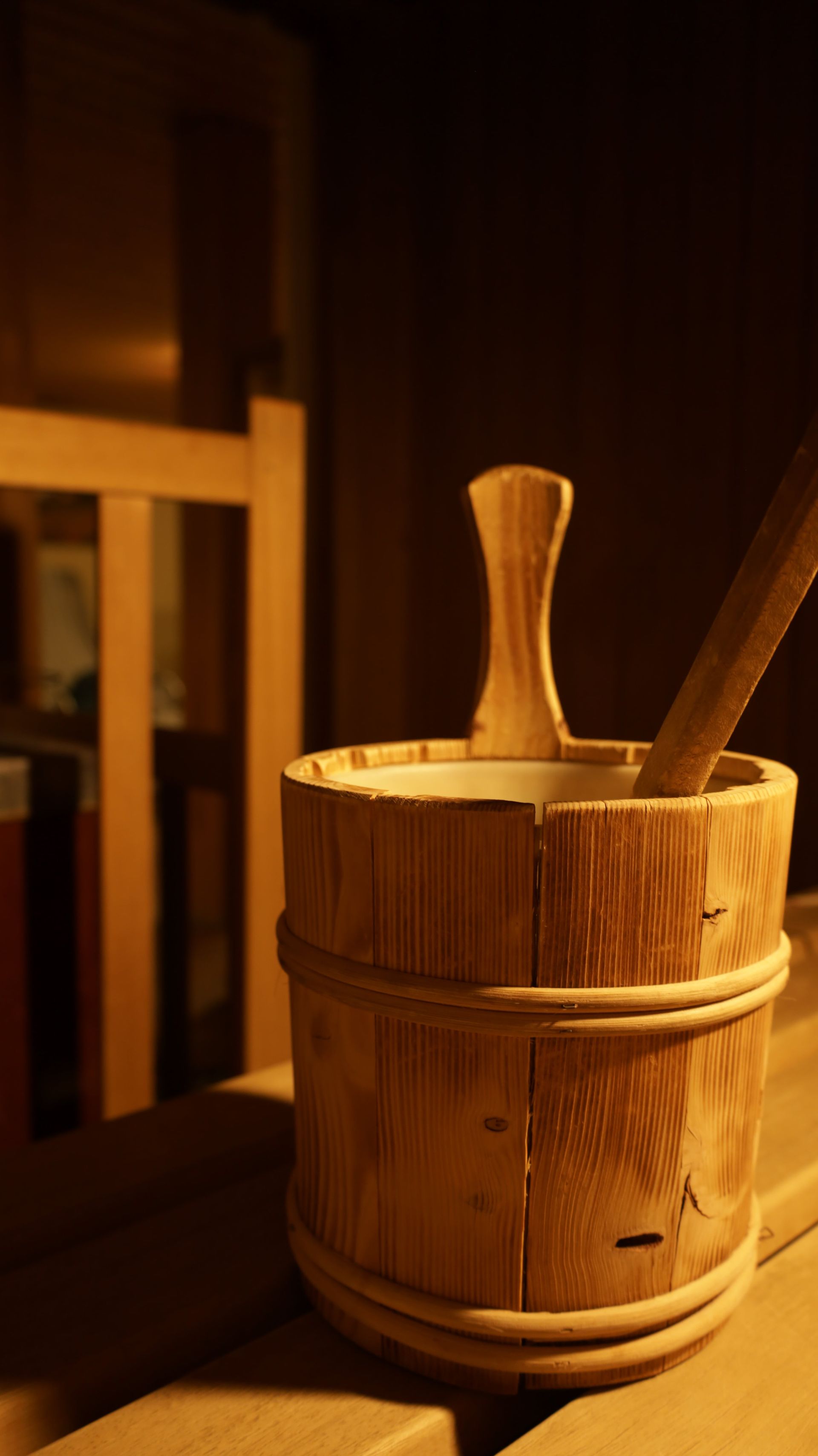 Ein Holzeimer mit einem Holzlöffel darin steht auf einer Holzbank in einer Sauna.