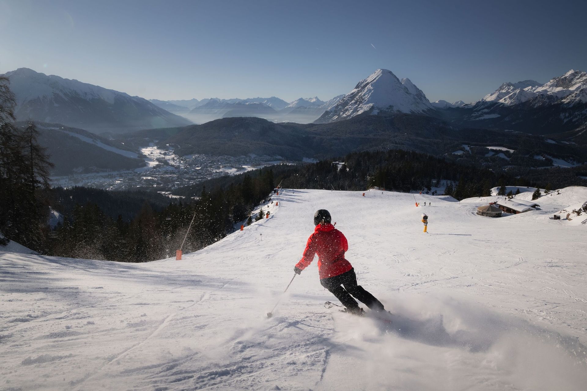 Eine Person fährt mit Skiern einen schneebedeckten Hang hinunter, im Hintergrund sind Berge zu sehen.