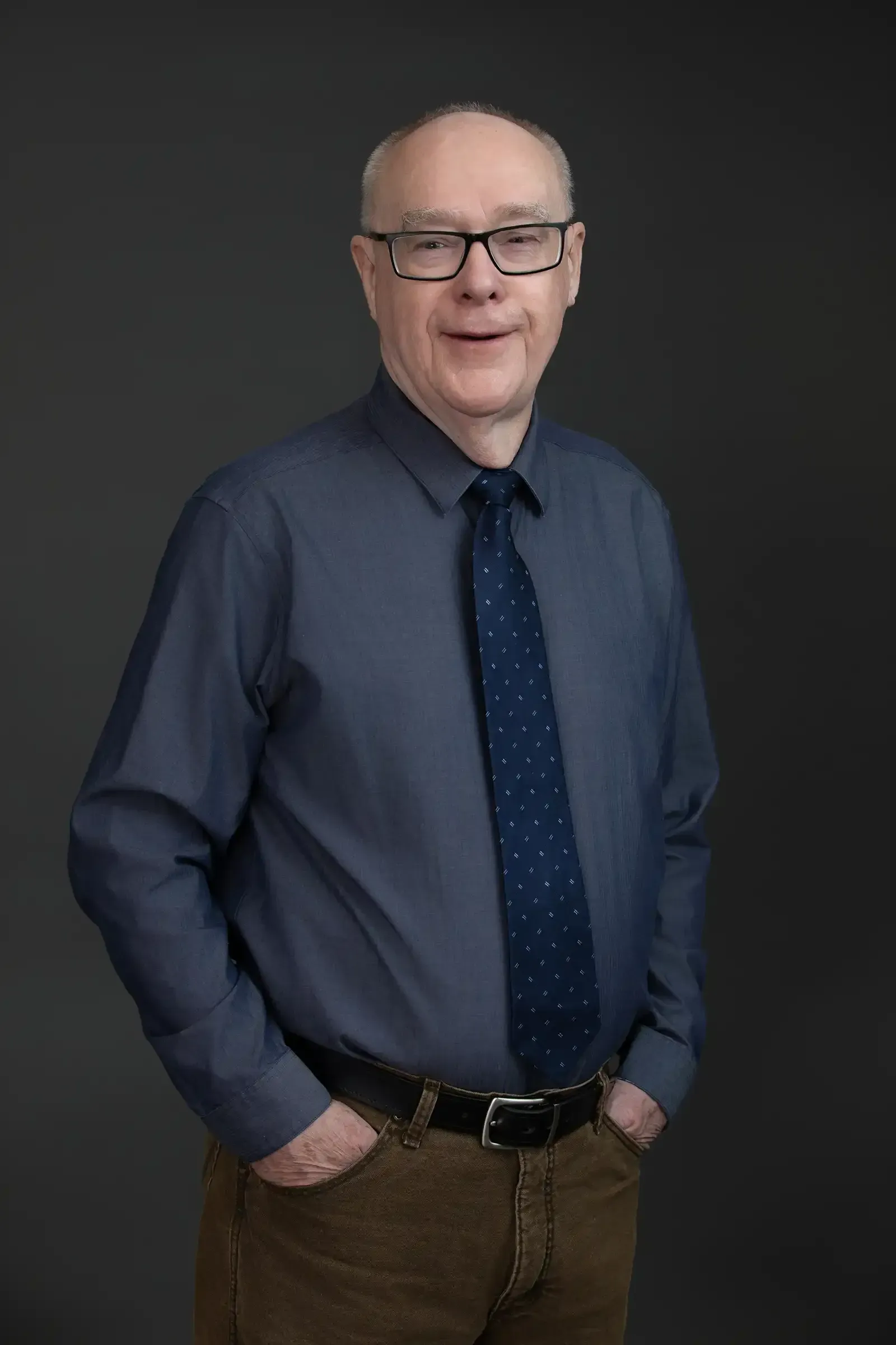 Chuck McClure in a blue shirt and tie is standing with his hands in his pockets.