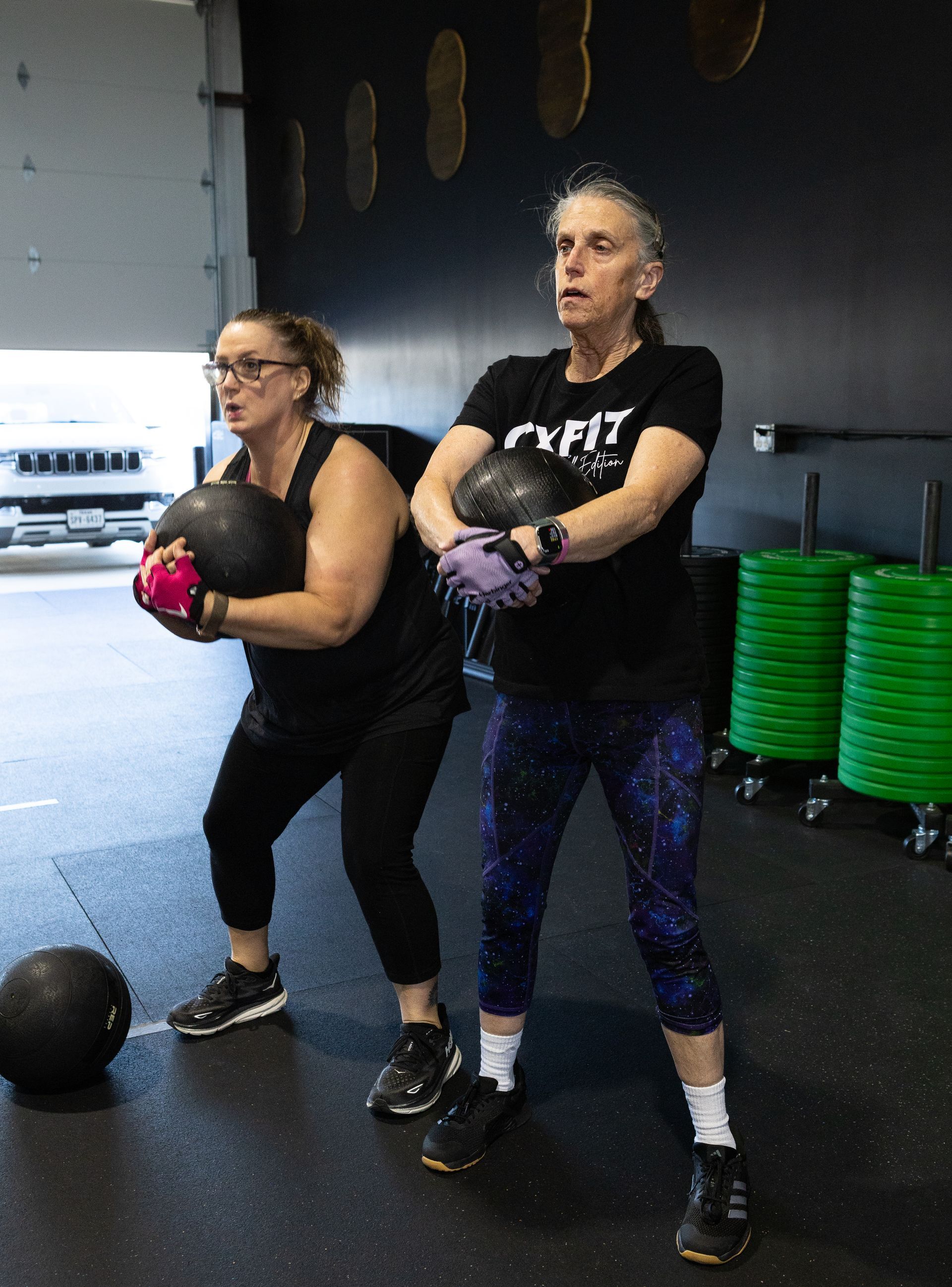 Two people holding medicine balls in a gym, standing in workout clothes near stacked green step platforms.