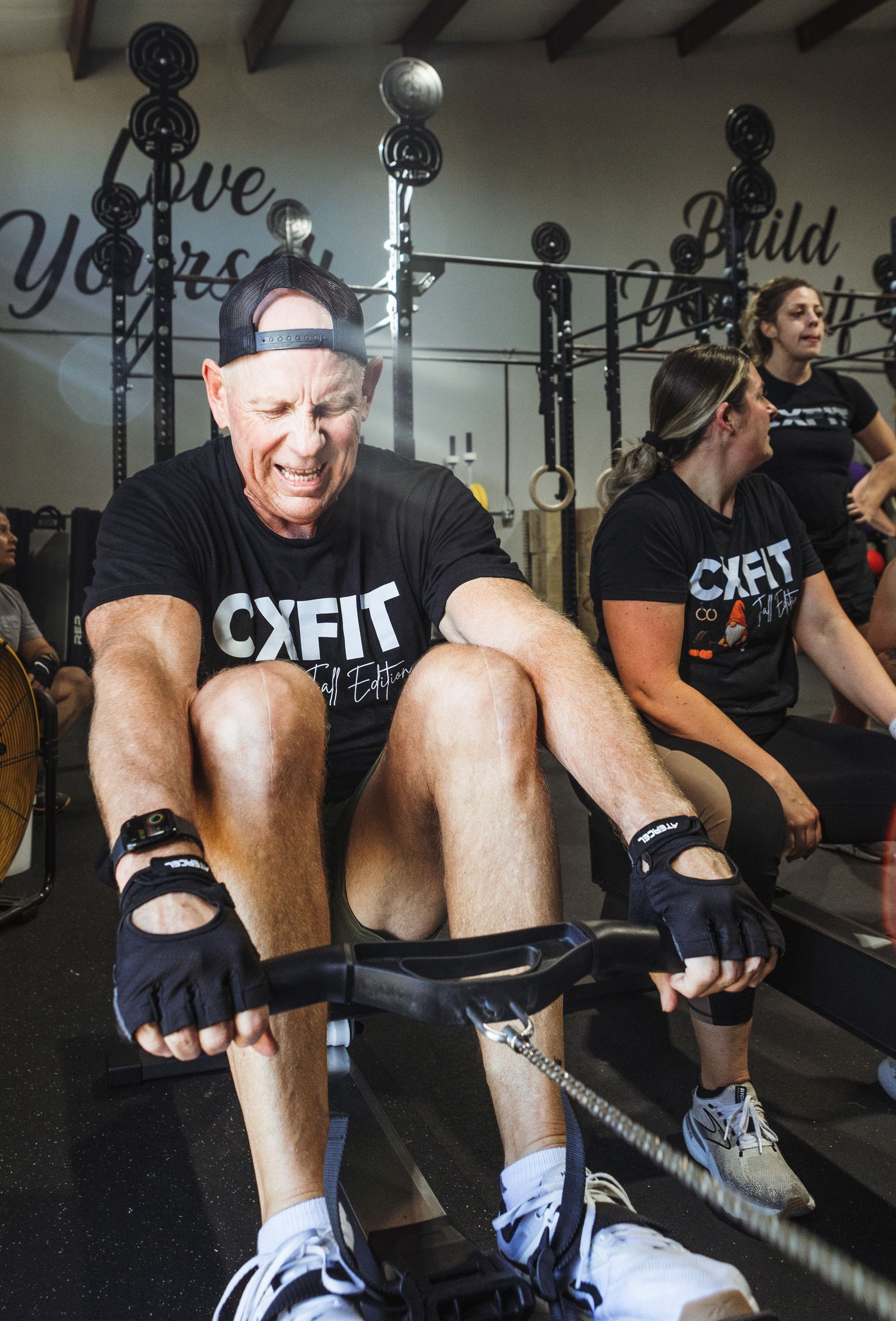 Athletes rowing on indoor machines in a gym, wearing black CXFIT shirts.