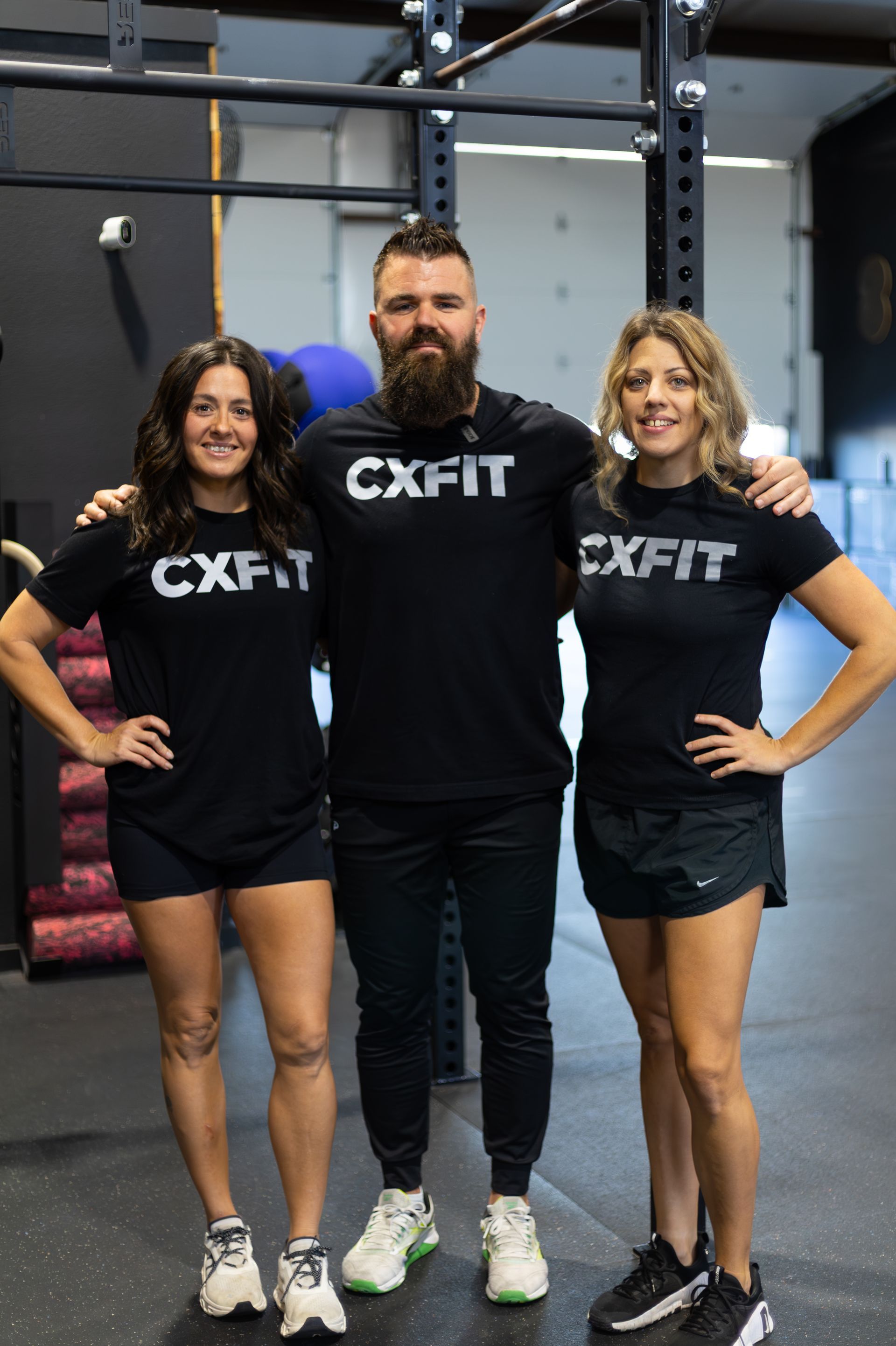 Three people in CXFIT shirts posing in a gym beside a squat rack.