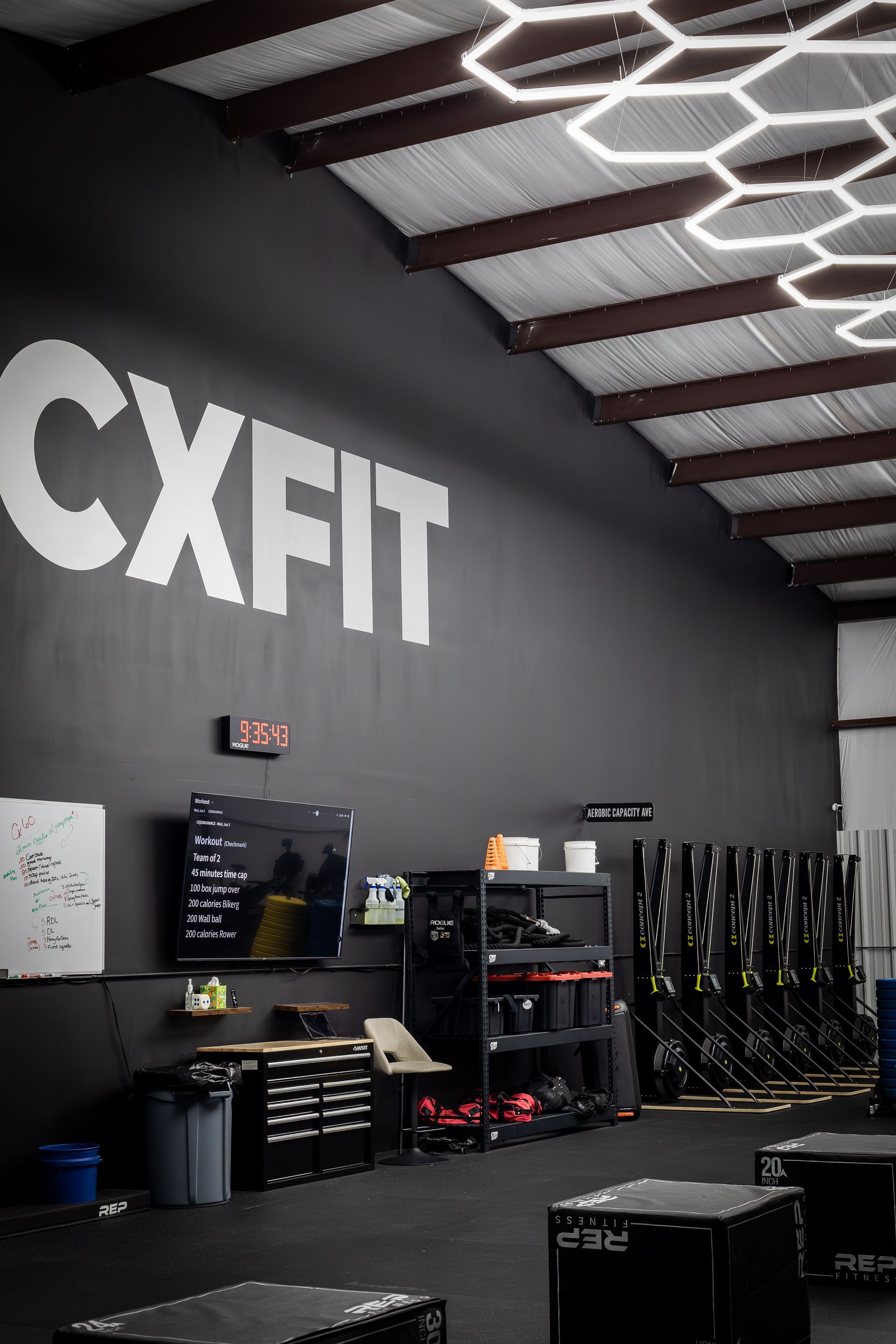 CXFIT gym interior with black wall, hex ceiling lights, and workout equipment along the side.