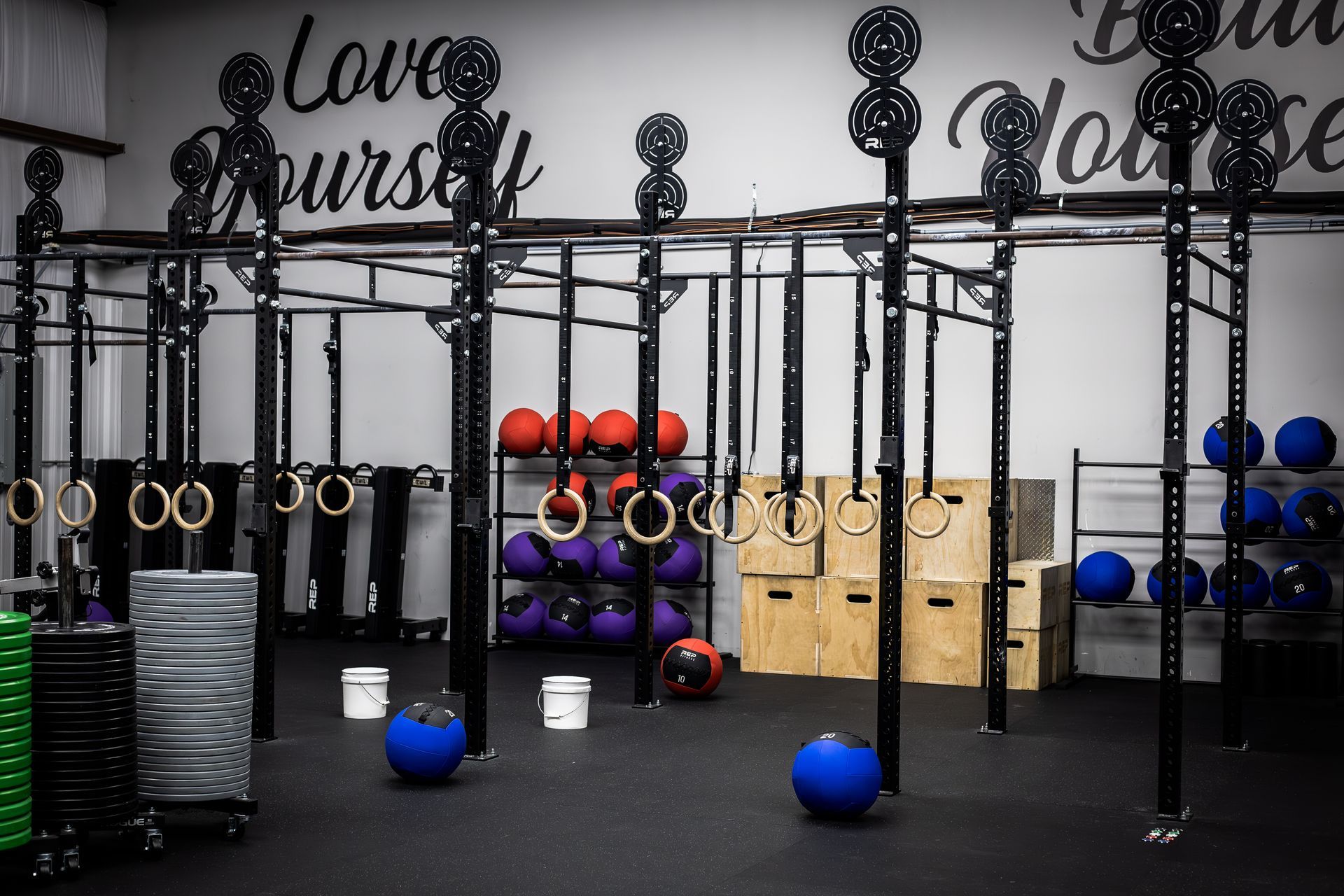 Gym with wall-mounted exercise rigs, kettlebells, medicine balls, and blue stability balls on a black floor.