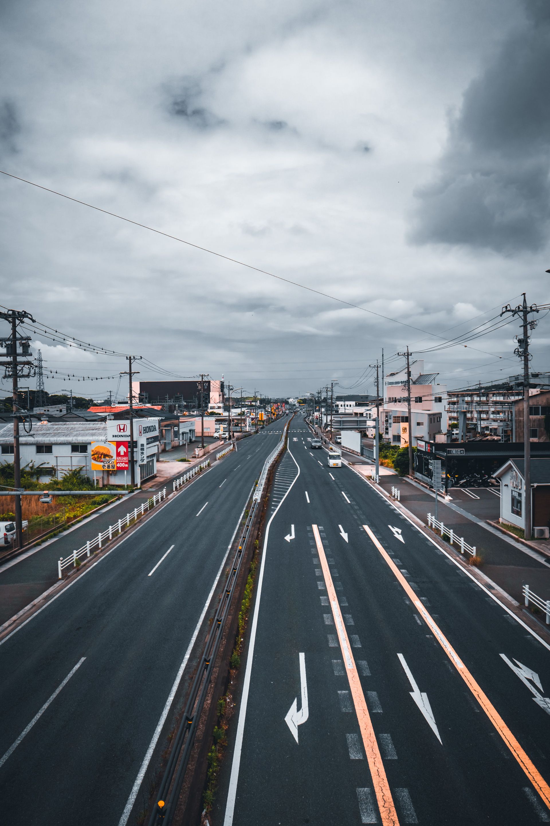 Empty multi-lane road stretching into a cloudy sky, with buildings and utility poles on both sides.