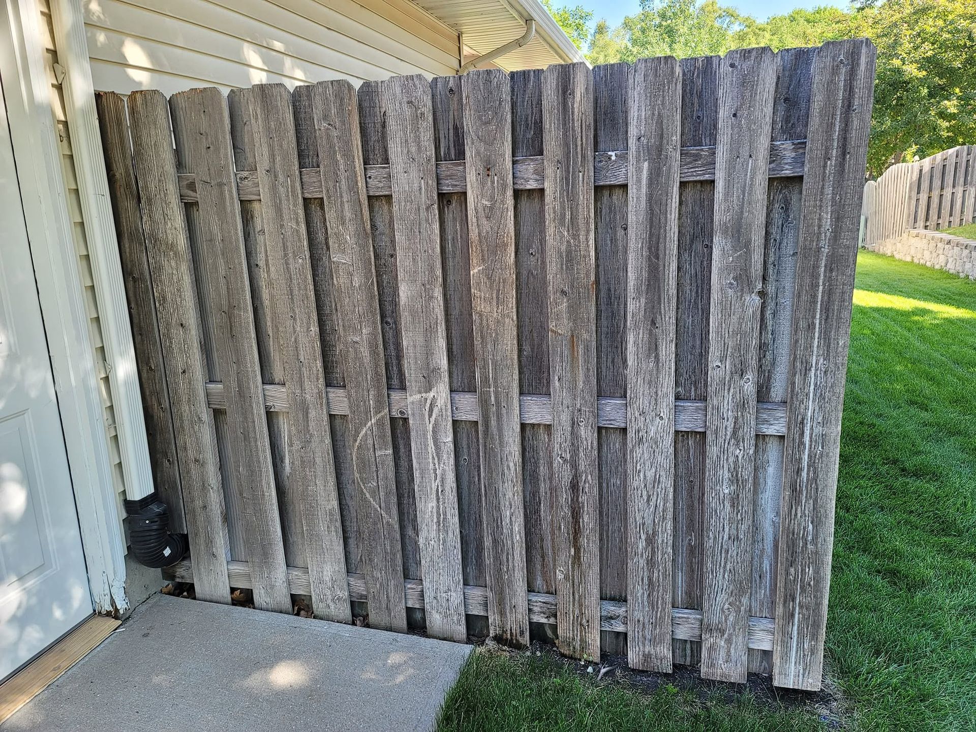 Weathered wooden privacy fence beside a white house, partly blocking a grassy yard.