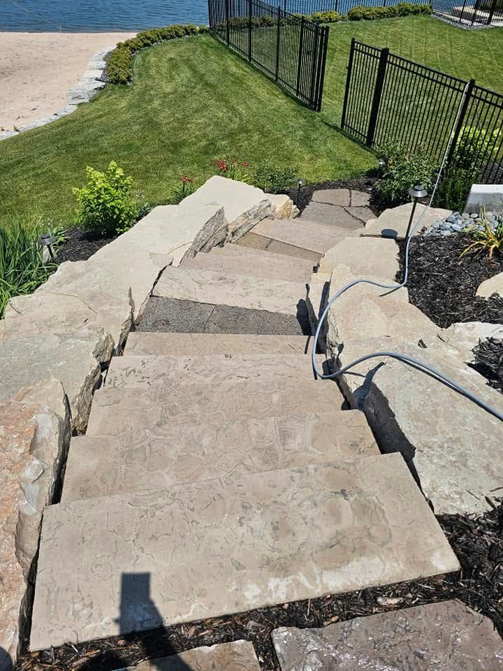 Stone steps descending a grassy slope beside a black fence toward a sandy shoreline