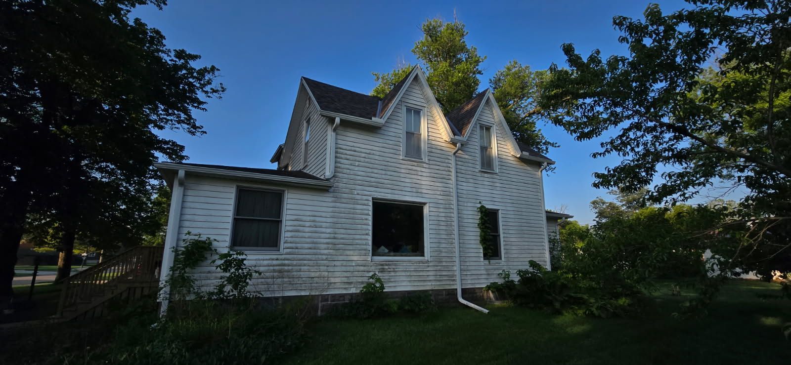 Old white house with peeling paint and gabled roof, partly shadowed by trees under a blue sky.