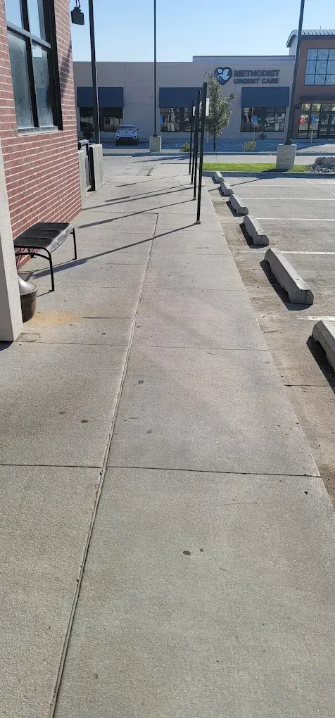 Sidewalk beside a brick building, with parking lot and storefronts in the distance.