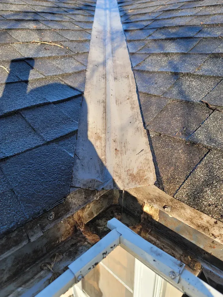Damaged roof ridge with peeling white flashing between dark gray shingles over a corner window