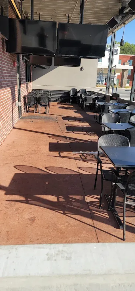 Covered outdoor patio with black tables and chairs on a red concrete floor, beside a brick storefront.