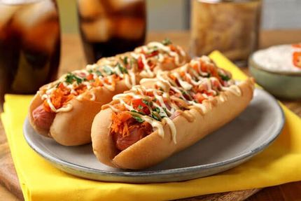Two hot dogs topped with sauce and herbs on a white plate, with a yellow napkin and drinks in the background