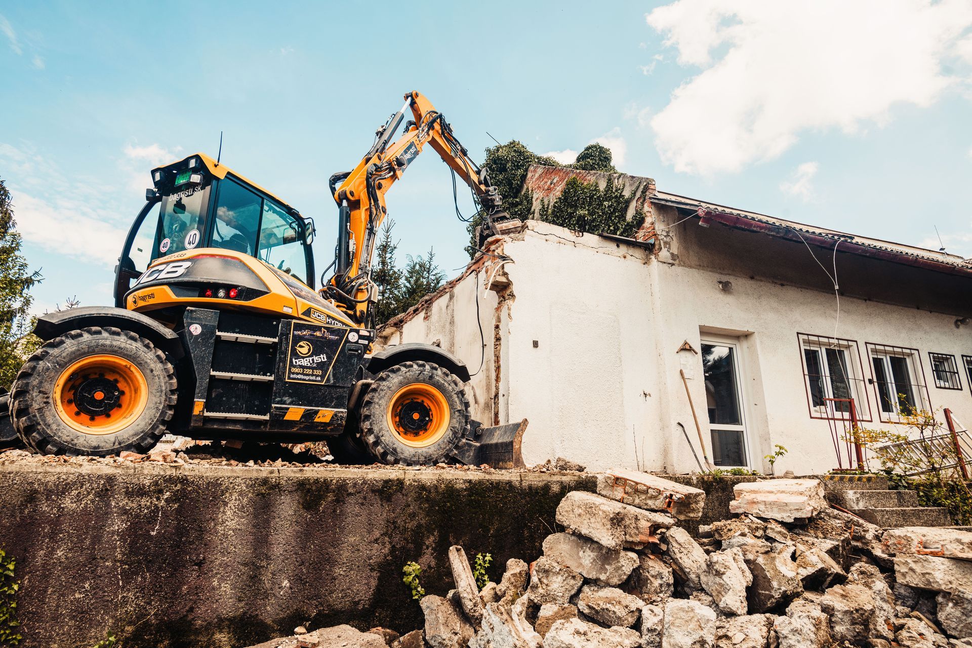 Excavator demolishing a damaged house, with rubble piled in the foreground.