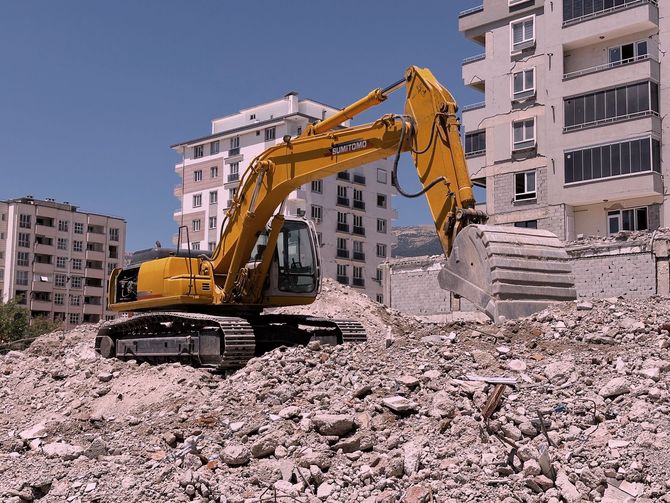 Yellow excavator on rubble amid damaged apartment buildings at a demolition site