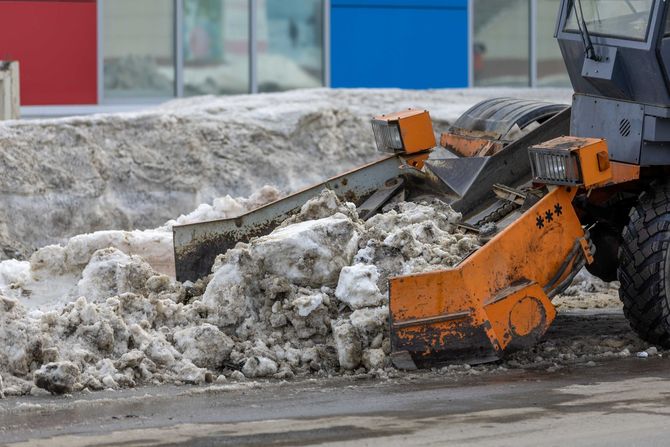 Orange snowplow clearing dirty snow pile along a city street beside a red and blue building