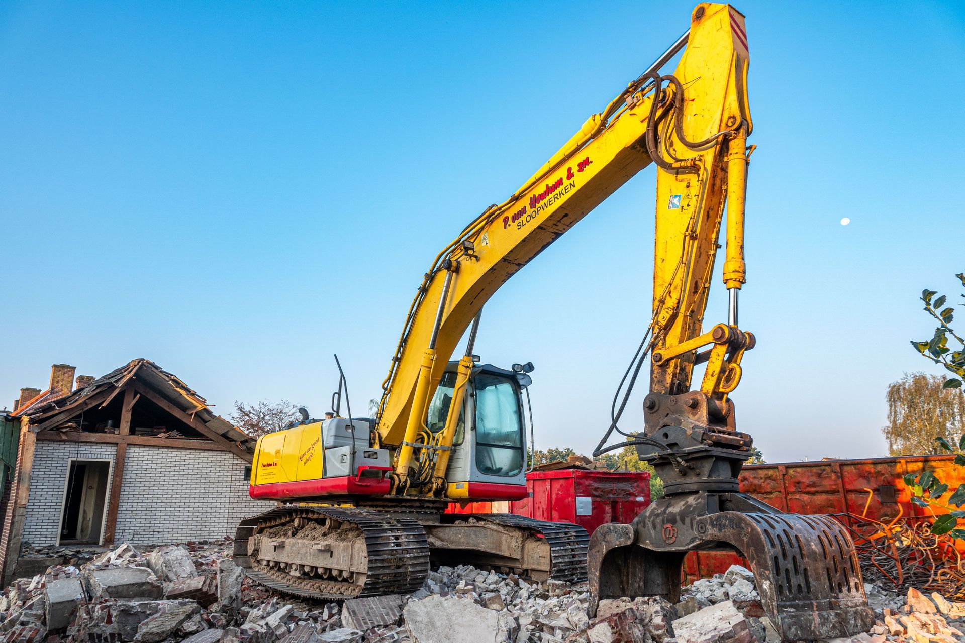 Yellow excavator demolishing rubble beside a damaged building under a blue sky