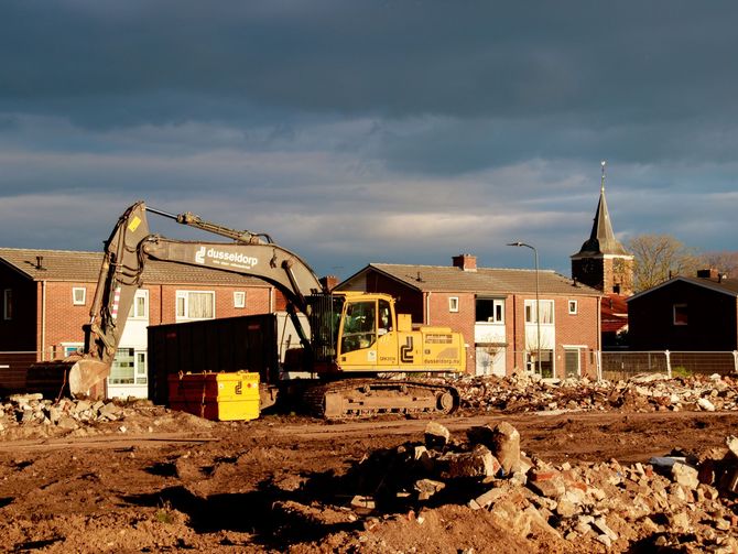 Excavator clearing rubble at a demolished building site with brick houses in the background.