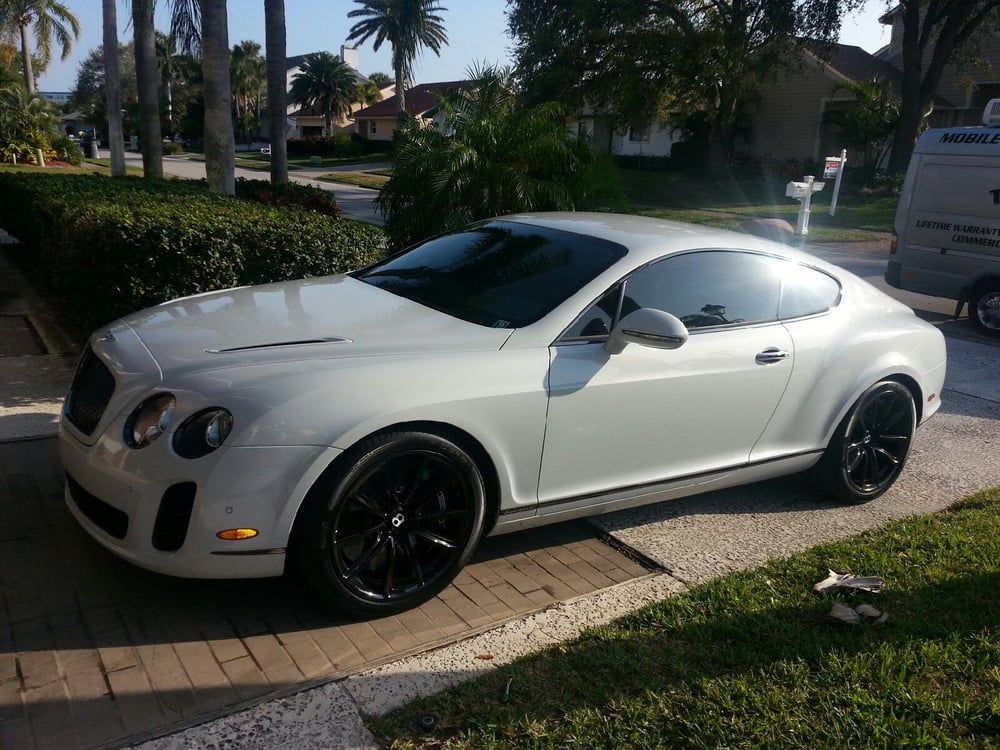 White Bentley coupe parked on a driveway with black wheels, sunny day.