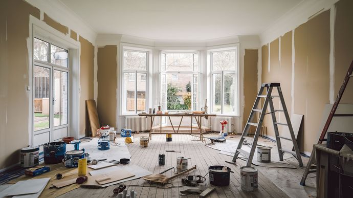 Pièce en rénovation avec des pots de peinture, des outils et des échelles ; fenêtres en arrière-plan.