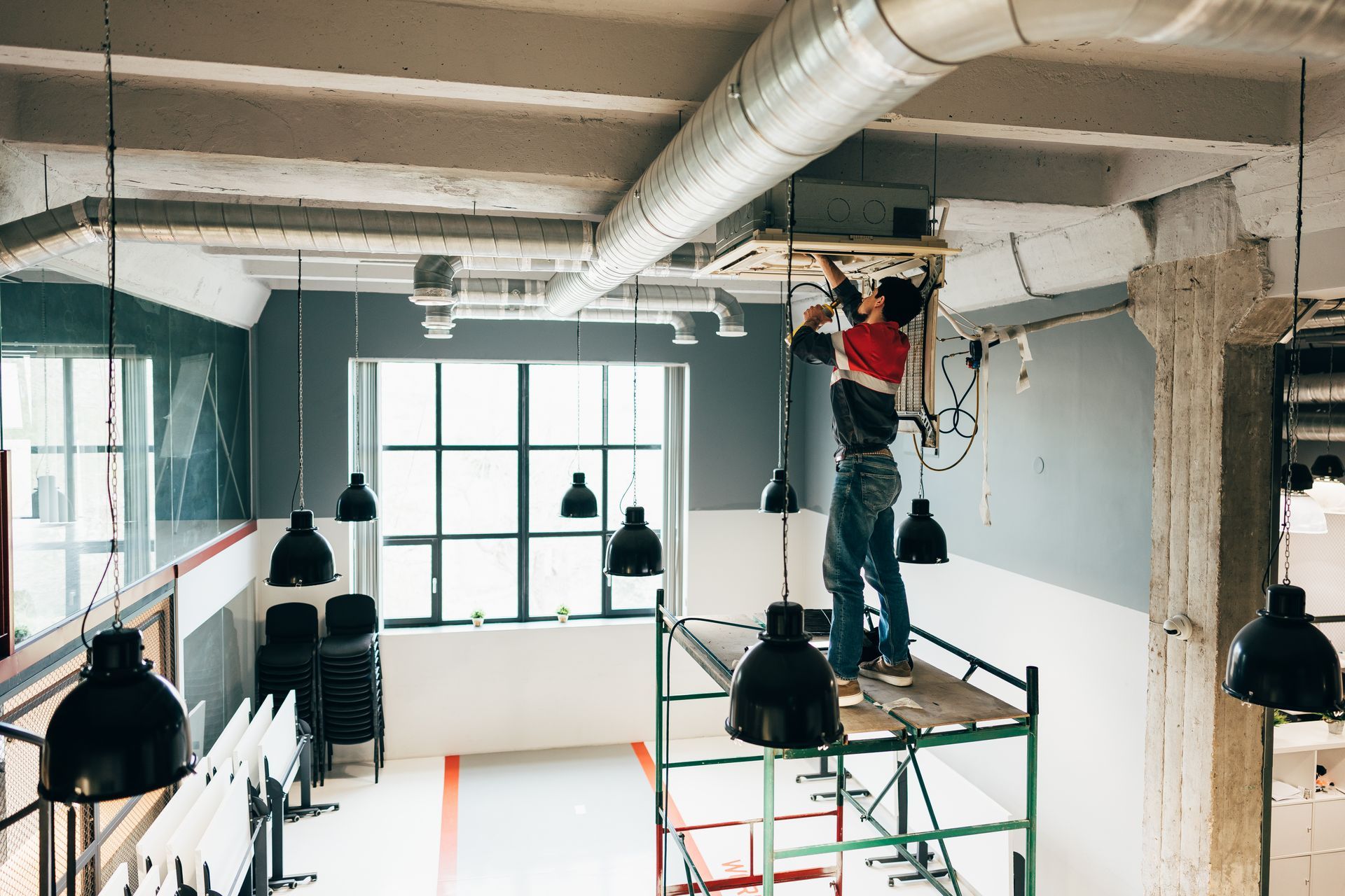Une personne sur un échafaudage installe un luminaire dans un bâtiment de style industriel.