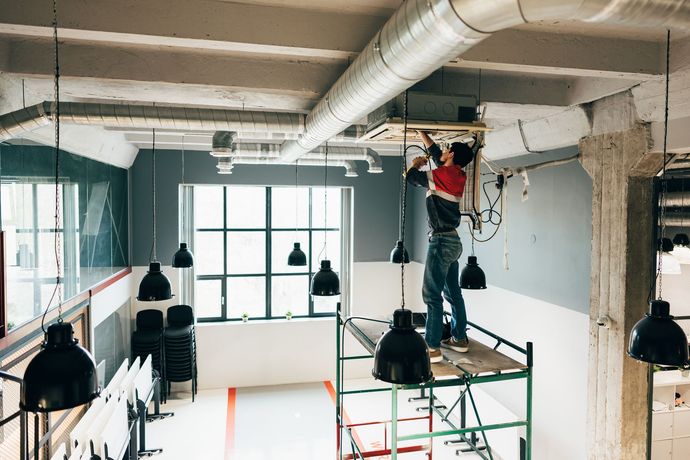 Une personne sur un échafaudage installe un luminaire dans un bâtiment de style industriel.