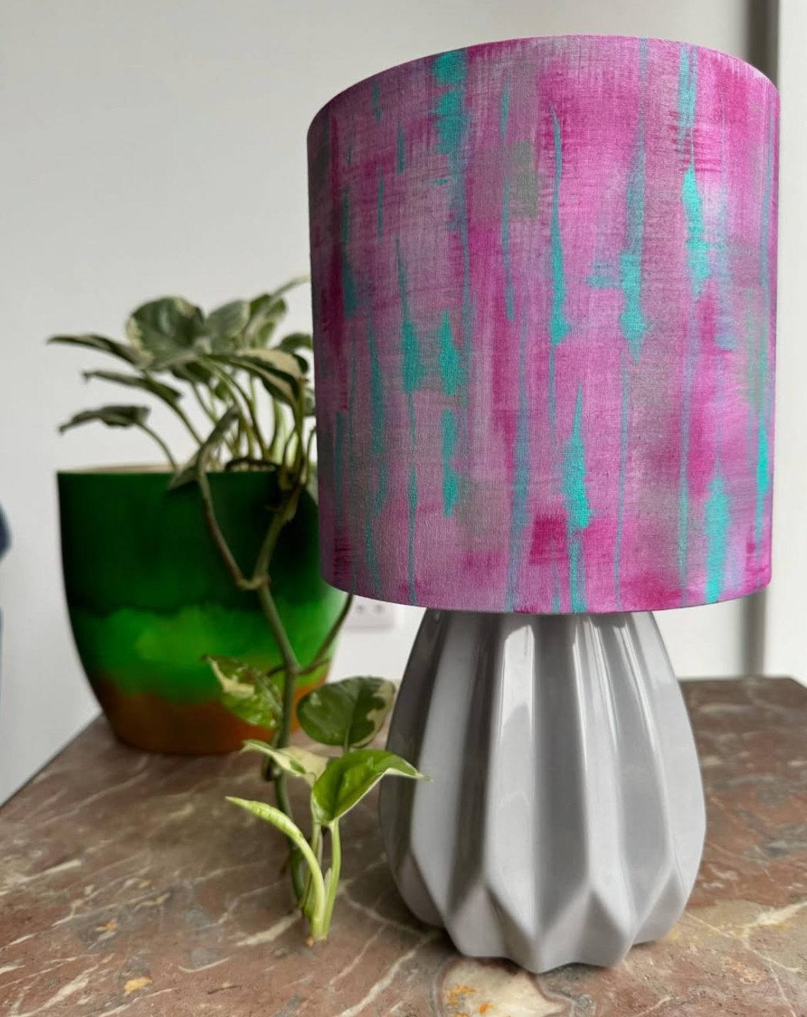 Table lamp with pink and teal shade, gray base, next to green plant in a green pot.
