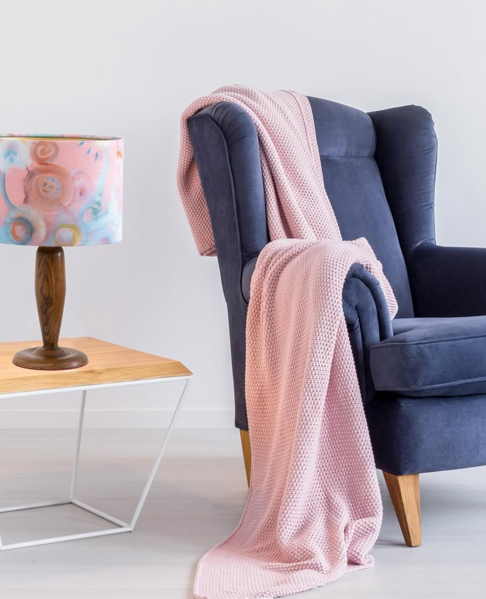 Blue armchair with pink blanket, next to a lamp on a table.
