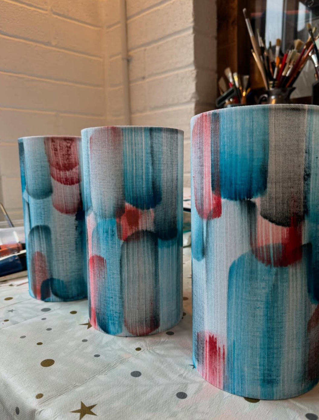 Three cylindrical lampshades with turquoise, red, and grey brushstroke patterns, sitting on a white tablecloth.