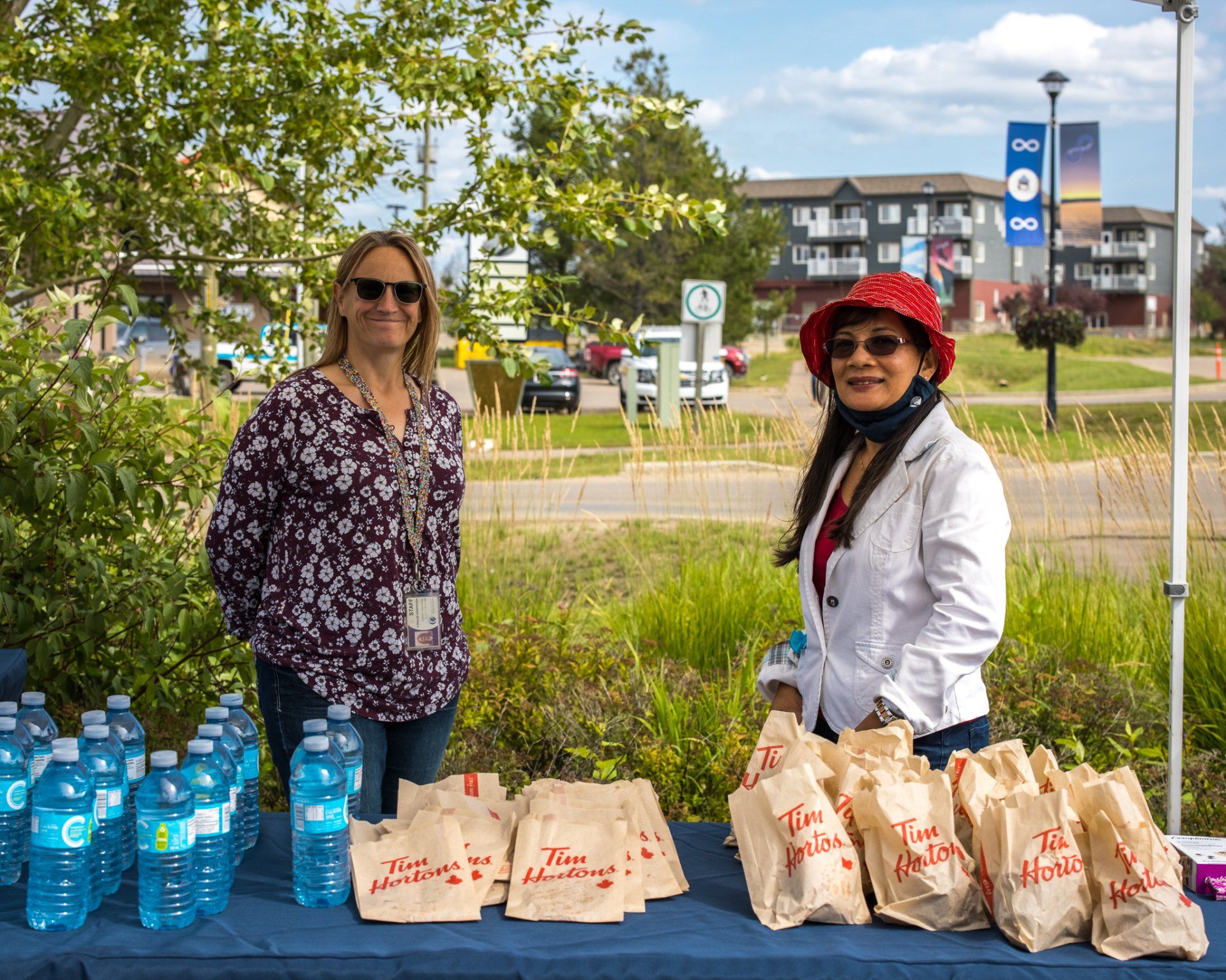 Two women stand behind a table with water bottles and Tim Hortons bags, outdoors.
