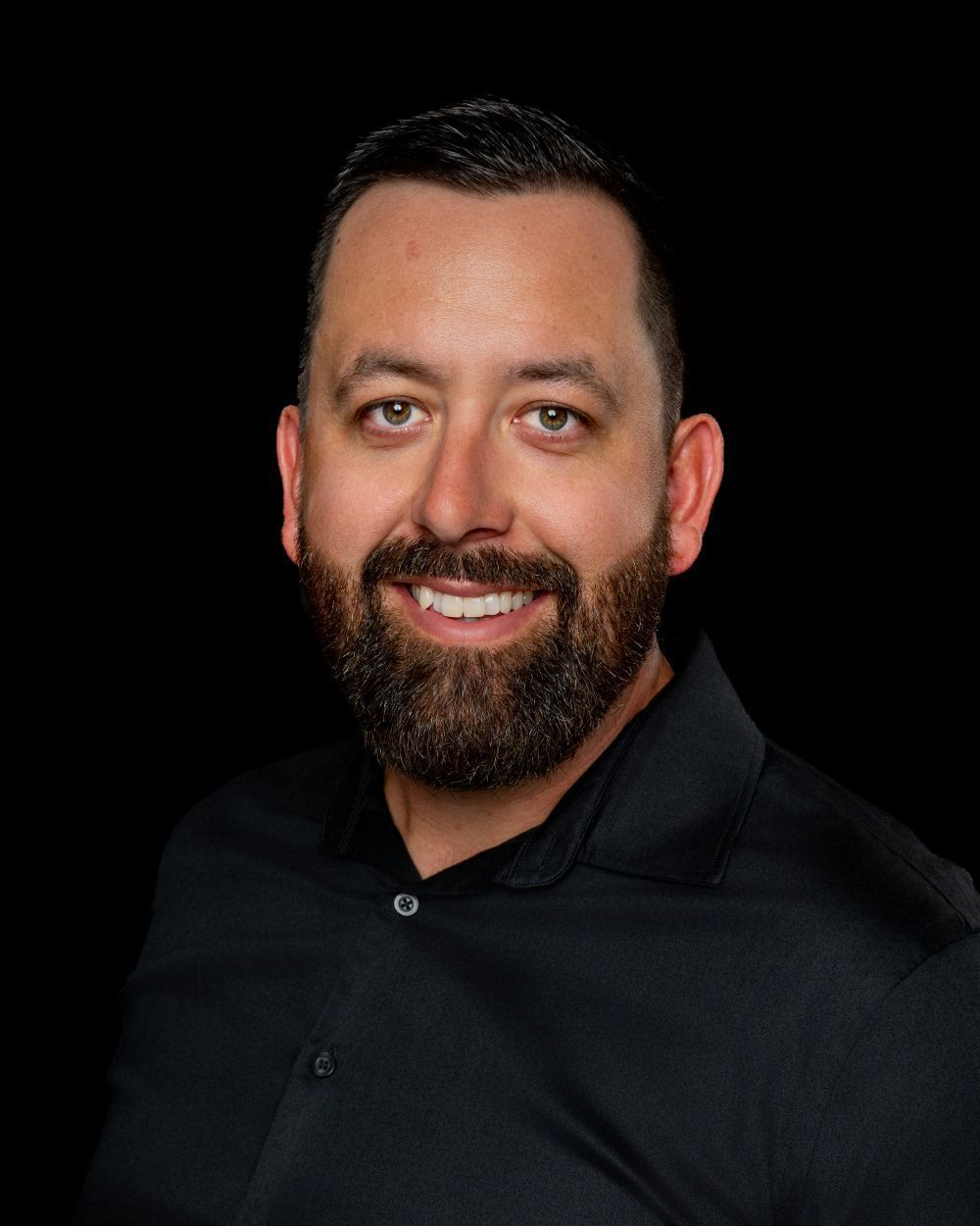 Smiling man with dark hair and beard wearing a black shirt against a black background