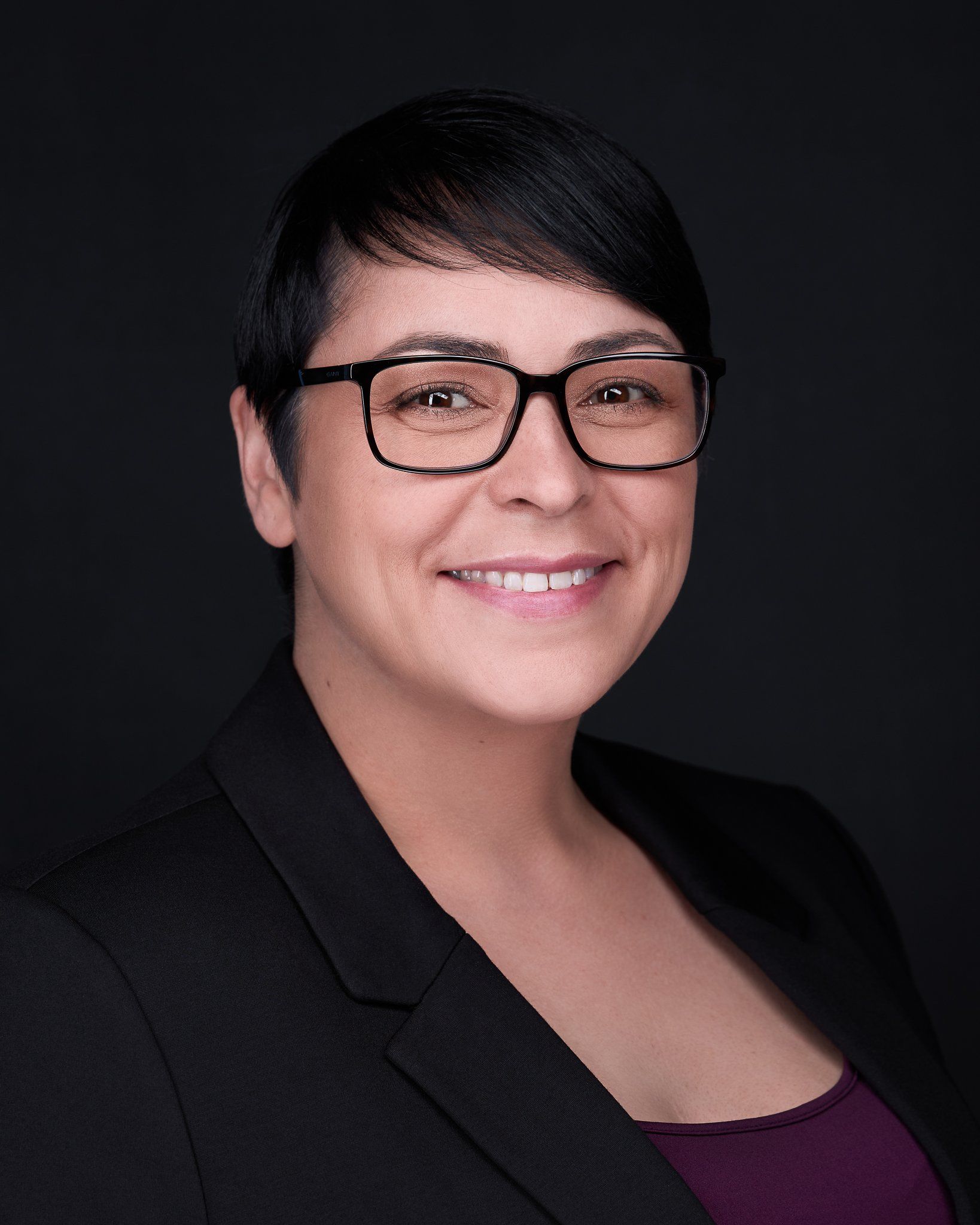 Woman with glasses smiles, wearing black blazer and purple top against a dark backdrop.
