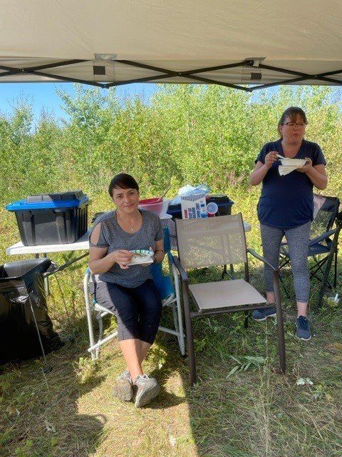 Two people outdoors under a canopy; one sits holding a snack, the other eats. A table with supplies and bushes are in the background.