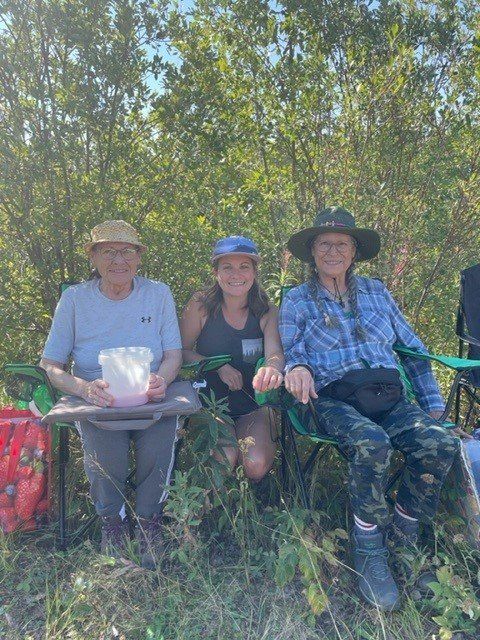 Three people seated in camping chairs outdoors, sunny day. One holds a container, another is kneeling. Green foliage in background.