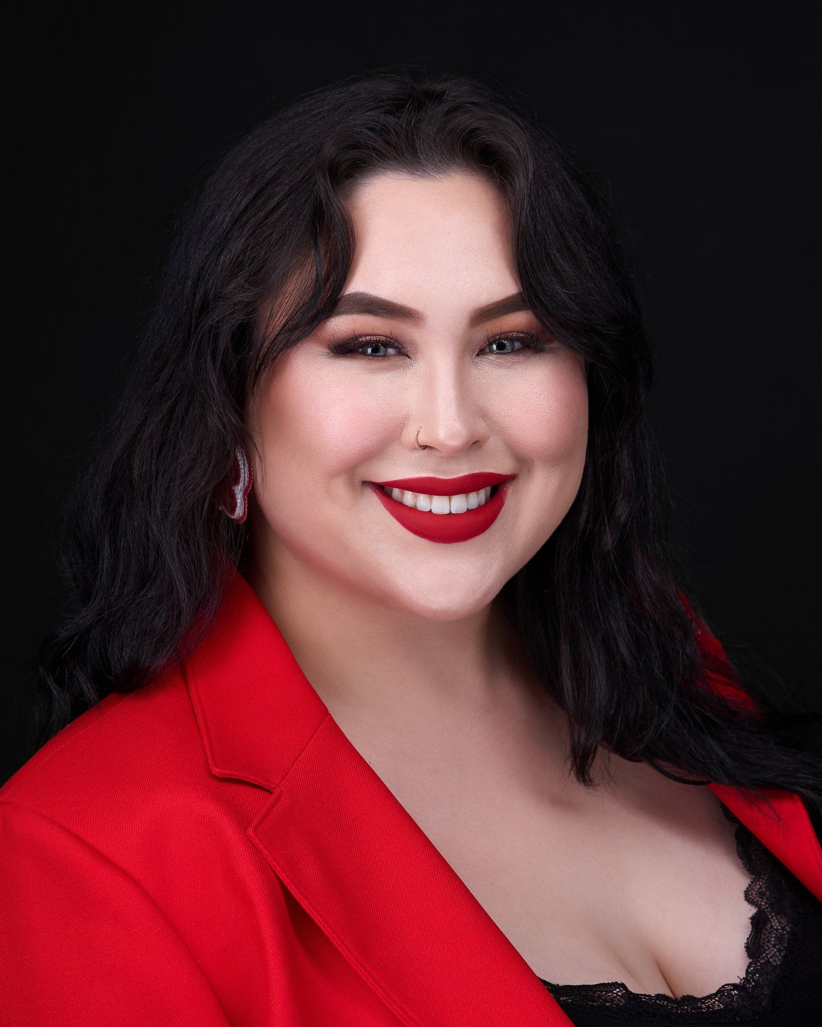 Woman in red blazer smiles at camera, dark hair, red lipstick, black background.