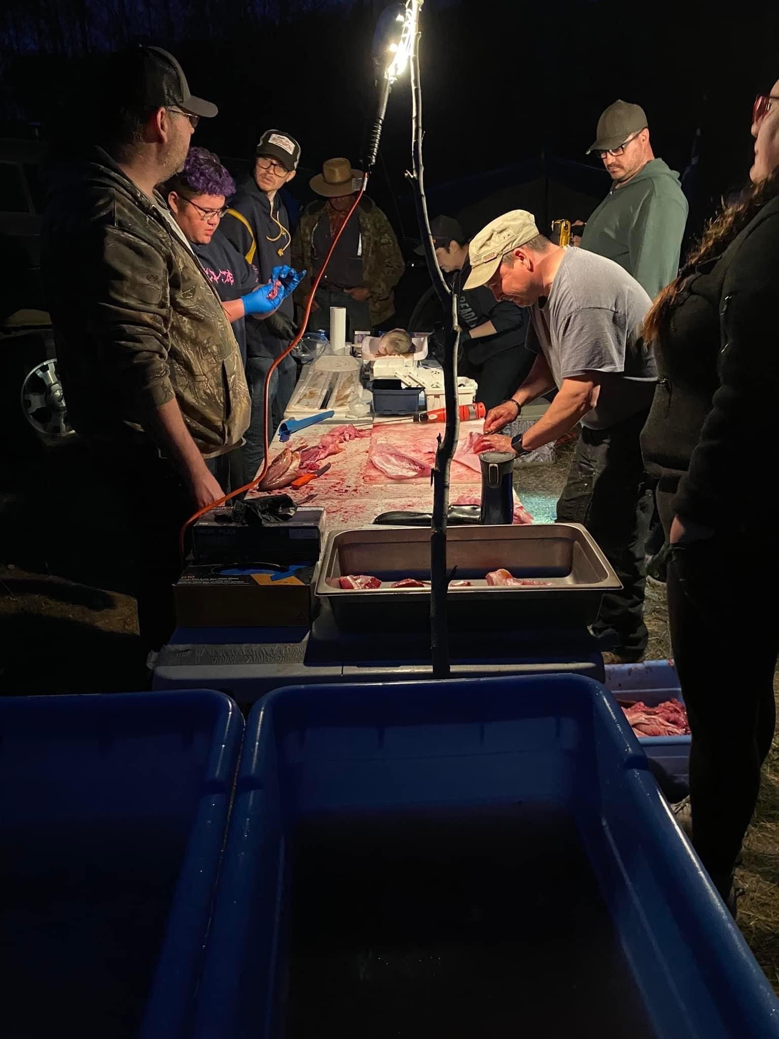 People gathered around a lit table, likely butchering an animal outdoors at night.