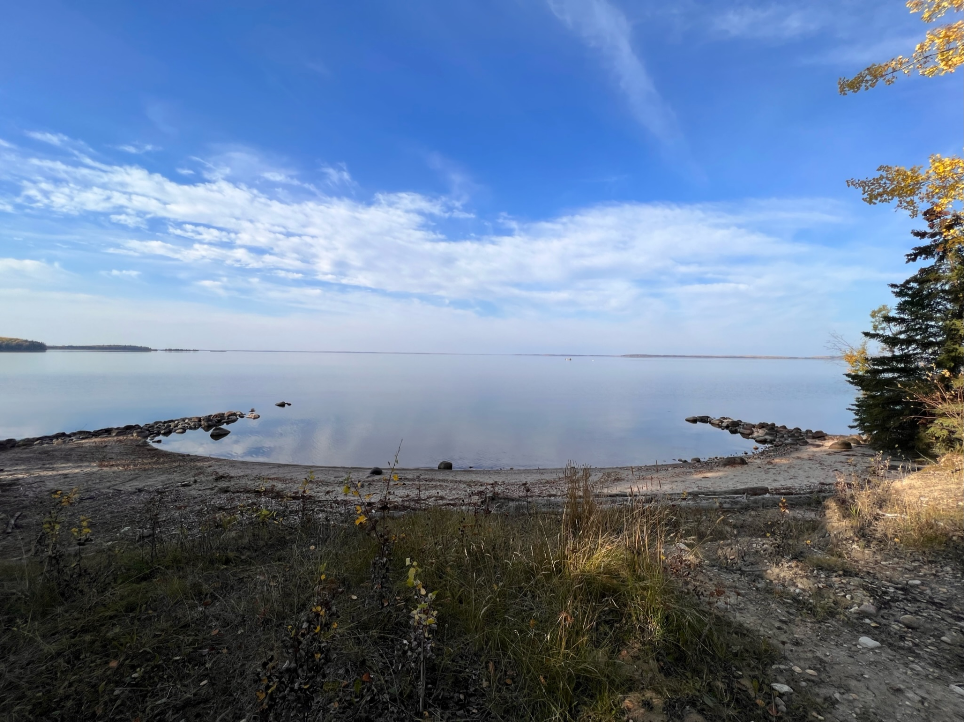 Calm lake view under a bright blue sky with scattered clouds. Rocks line the shore and trees on the right.