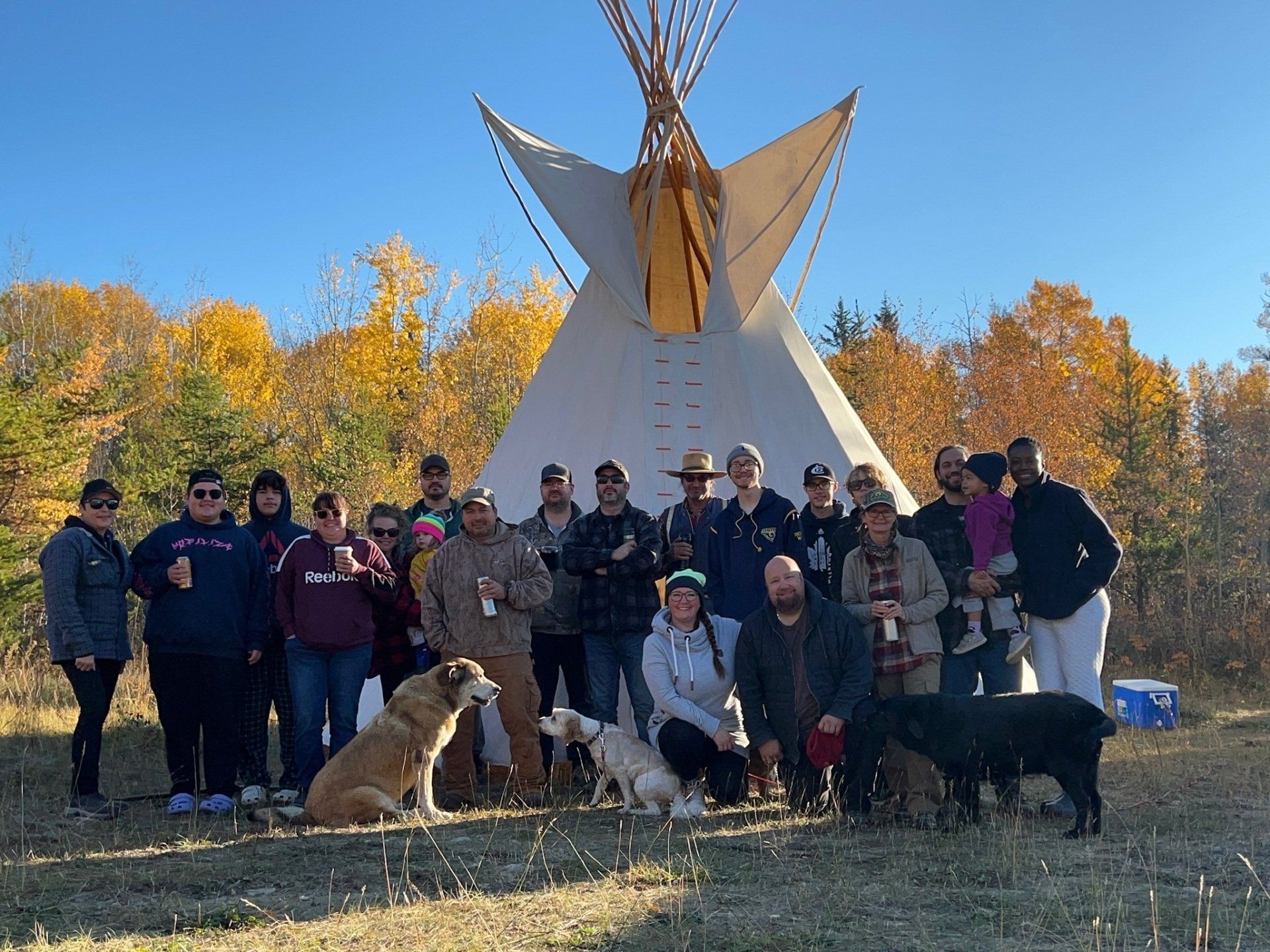 Group of people pose in front of a white teepee. Autumn trees in the background.