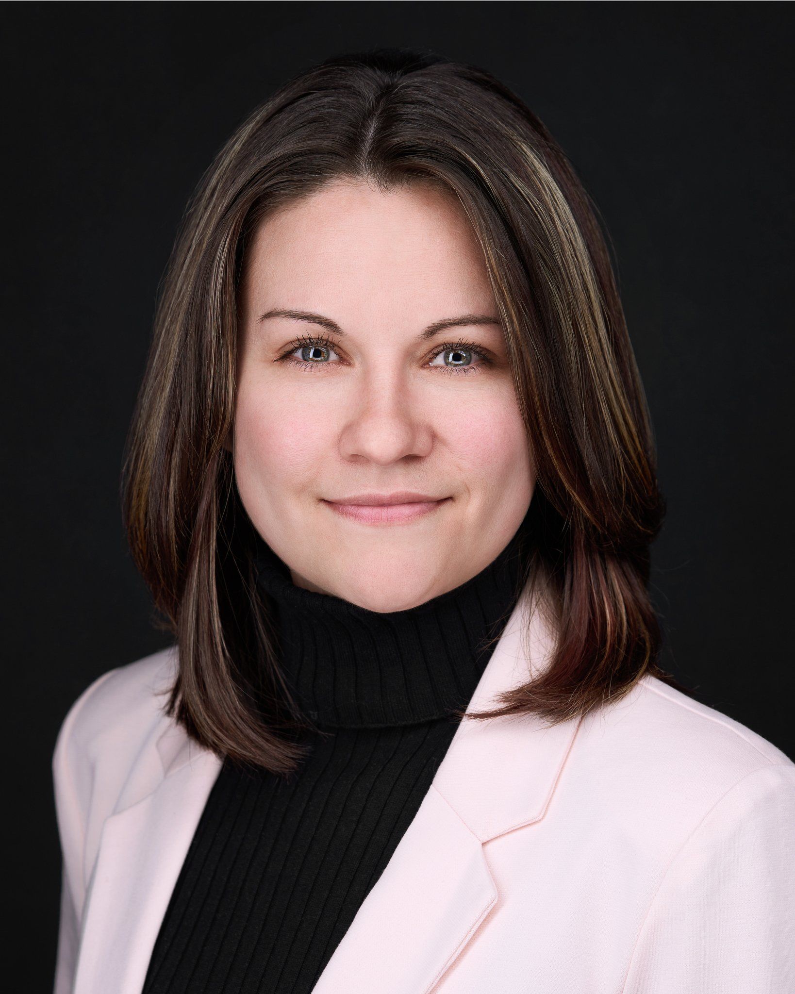 Woman in light pink blazer and black turtleneck, looking at the viewer, dark background.