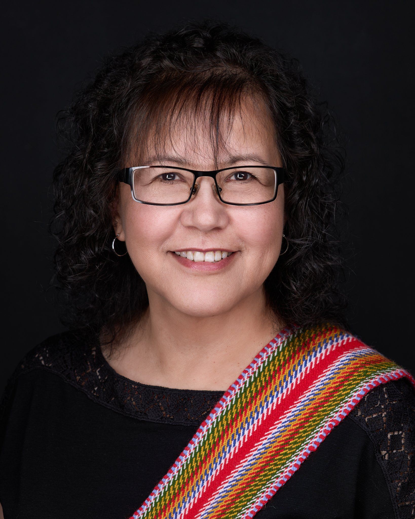 Woman with glasses smiles, wearing black top and black background.