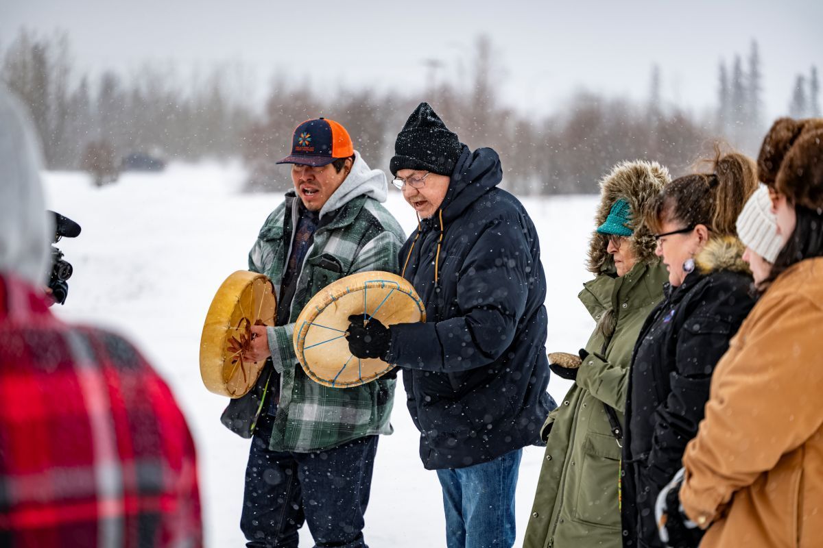 People in winter gear holding traditional drums outdoors in a snowy landscape.