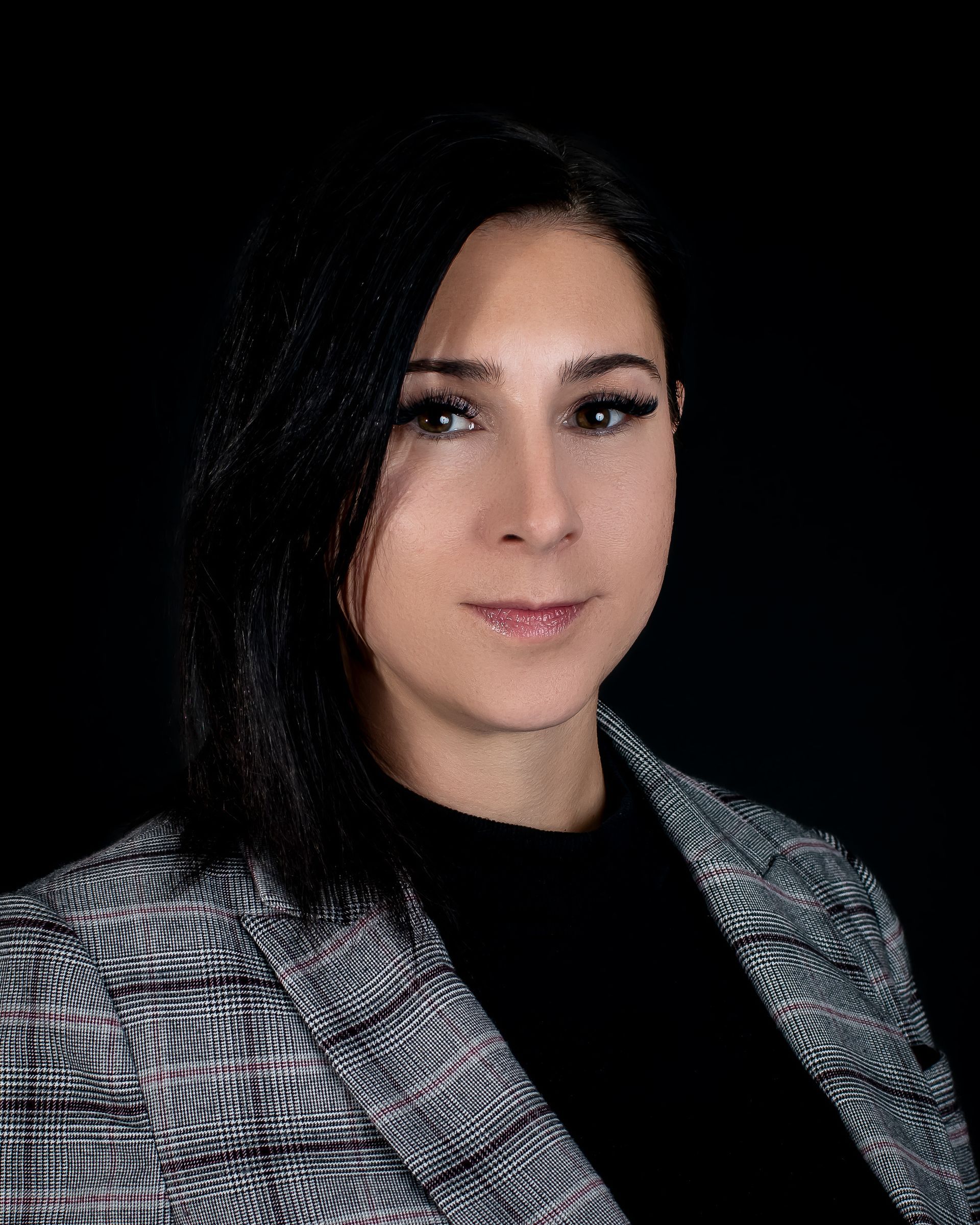 Woman with dark hair in a gray and black patterned blazer, black shirt against a black background.