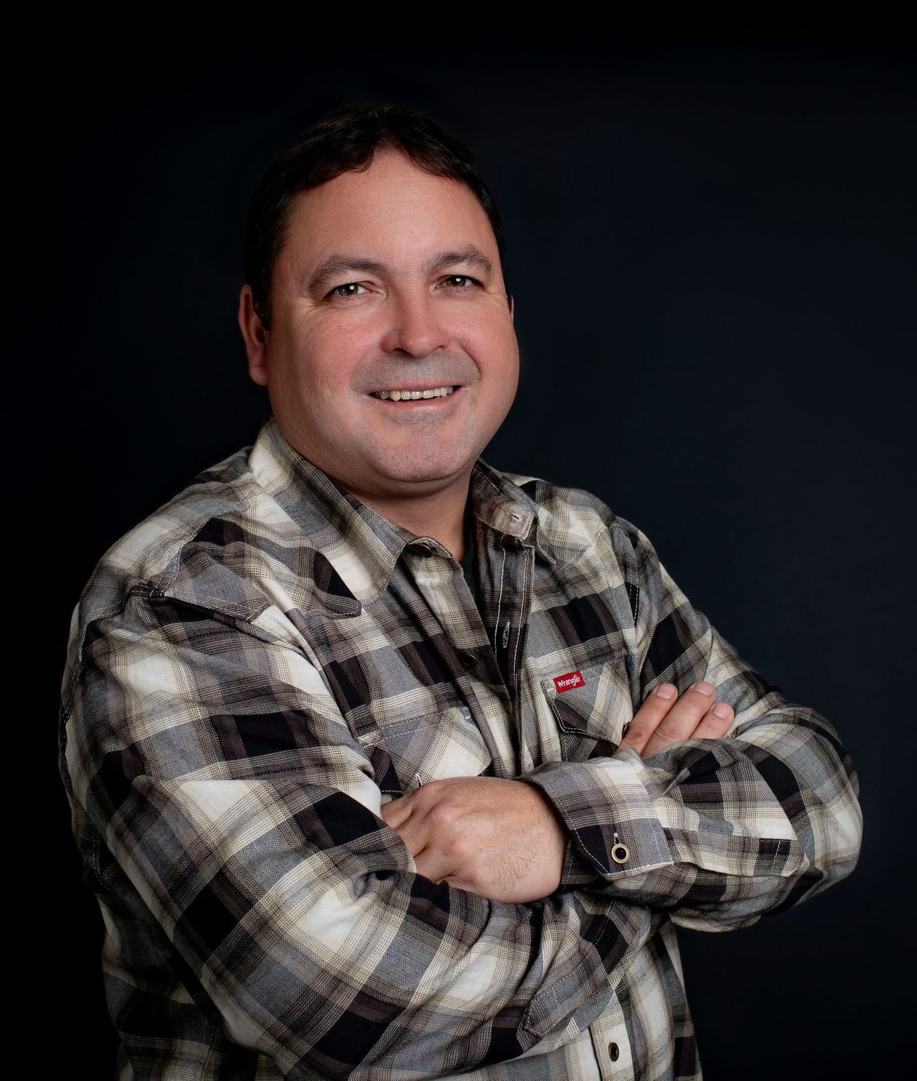 Man with arms crossed, wearing a plaid shirt, smiling, against a dark blue background.