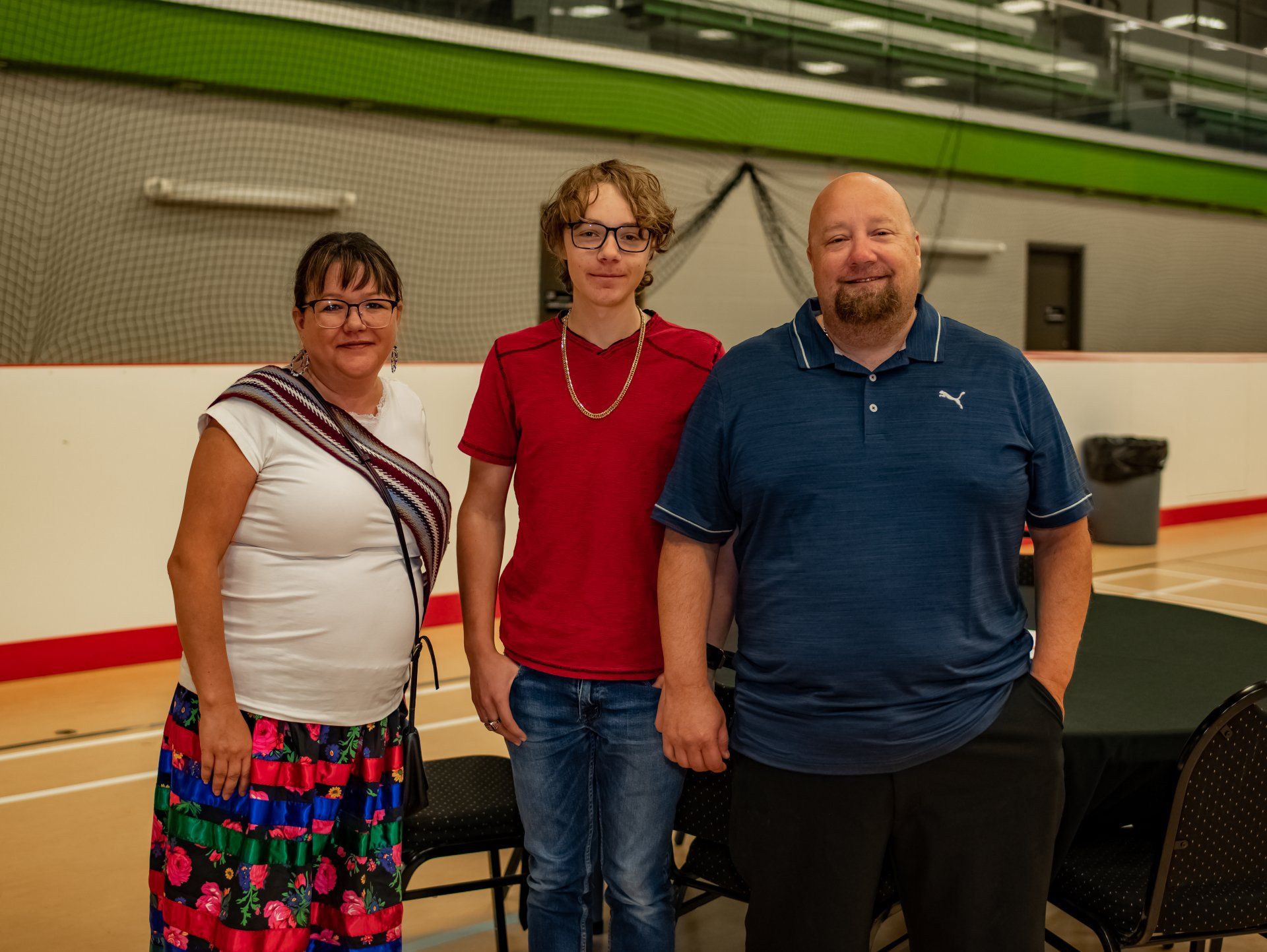 Three people stand indoors: woman in patterned skirt, teen in red shirt, man in blue shirt. Indoor setting.